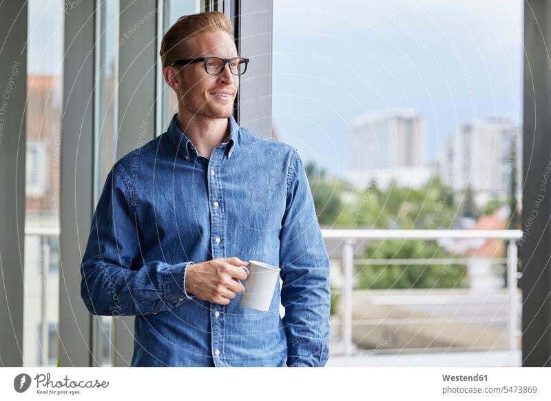 Lächelnder junger Mann mit Tasse Kaffee an der Balkontür stehend Kaffeetasse Kaffeetassen Balkontüren Balkontueren steht Männer männlich Tassen Getränk Getraenk