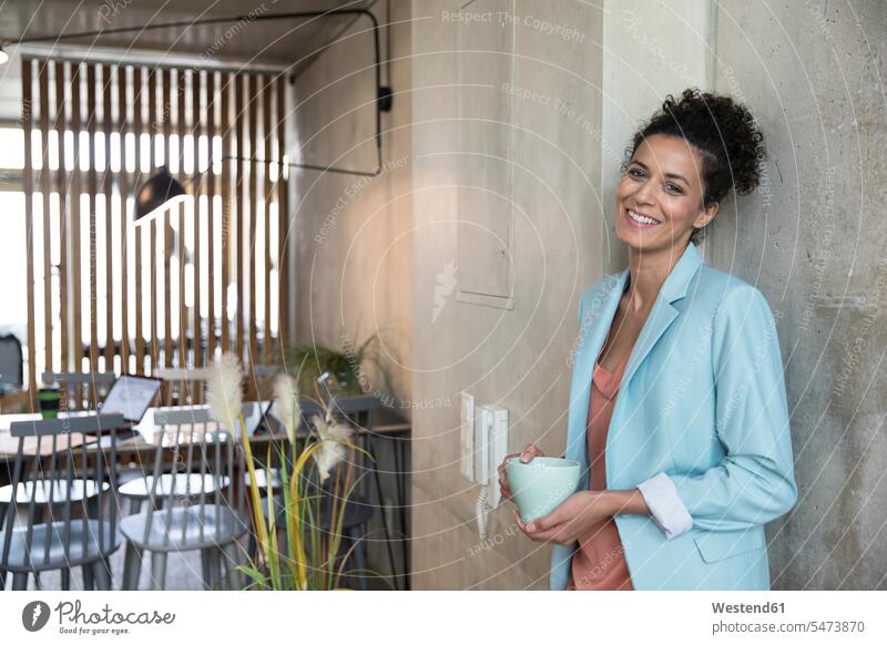 Porträt einer lächelnden Geschäftsfrau, die einen Kaffeebecher an einer Betonwand in einem Loft hält Betonwände Betonwaende Portrait Porträts Portraits Lofts