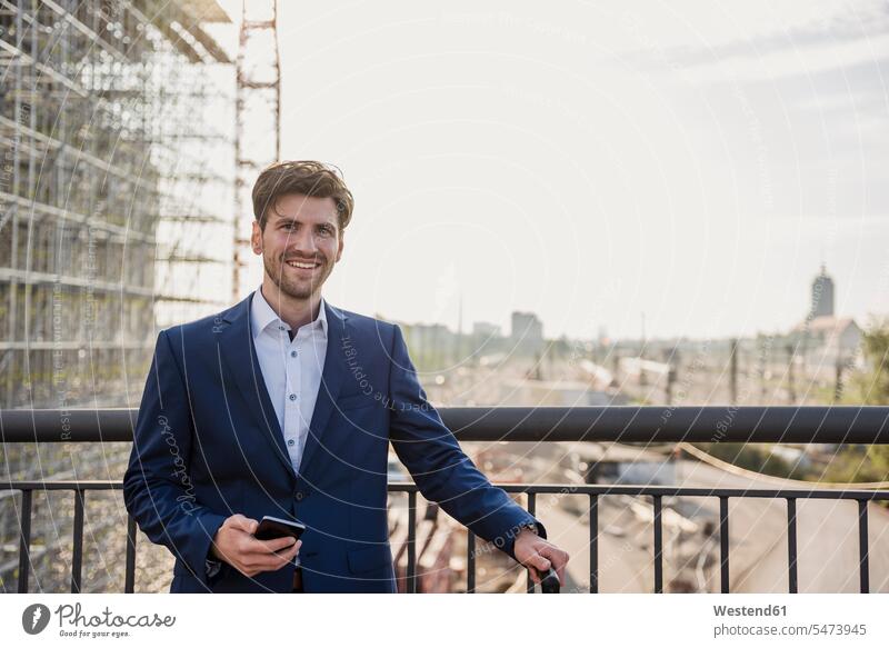 Porträt eines lächelnden Geschäftsmannes, der auf einer Brücke in der Stadt steht und ein Mobiltelefon hält Bruecken Brücken Businessmann Businessmänner