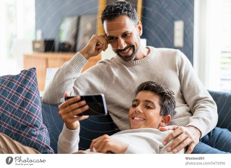 Lächelnder Vater und Sohn mit Smartphone auf der Couch im Wohnzimmer Couches Liege Sofas Telekommunikation telefonieren Handies Handys Mobiltelefon
