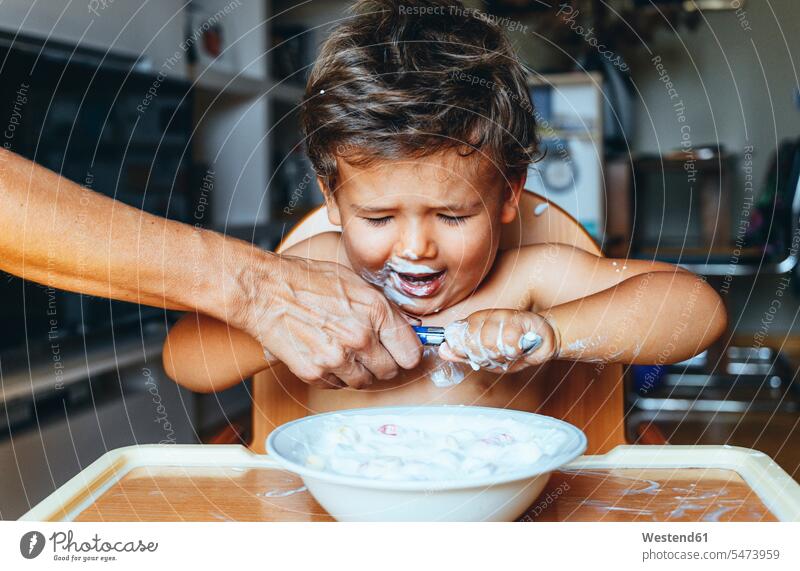 Kleiner Junge isst zu Hause Joghurt, Hand der Frau auf dem Löffel Schale Schalen Schälchen Schüsseln Loeffel Trotz Stütze stützen unterstützen gefühlvoll