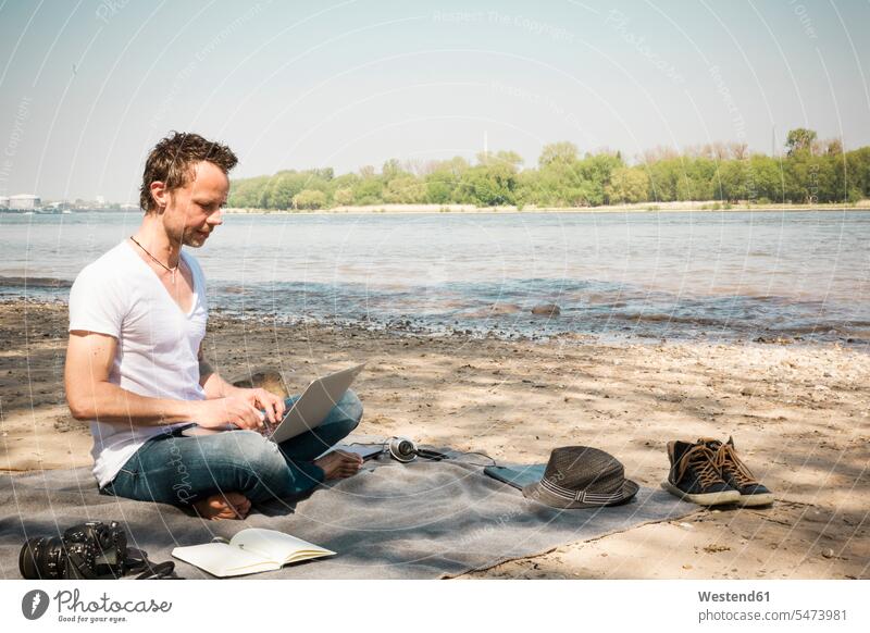 Mann sitzt mit Laptop auf einer Decke an einem Fluss Notebook Laptops Notebooks sitzen sitzend Männer männlich Decken Strand Beach Straende Strände Beaches
