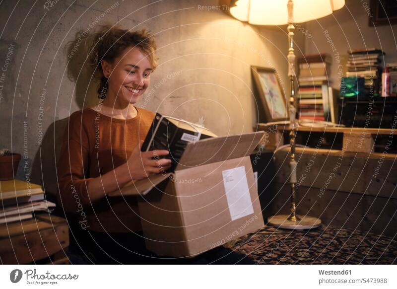 Lächelnde junge Frau packt Paket zu Hause aus Europäer eine Person 25-30 Jahre Licht Schatten Deutschland Innenaufnahme Freizeitkleidung auf dem Boden sitzen
