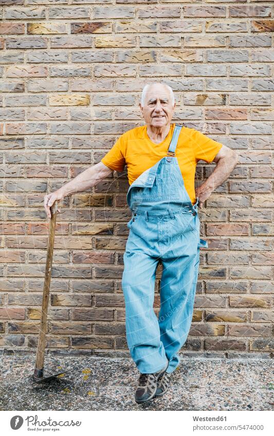 Älterer Mann in Jeans-Overall mit Gartengerät schaut in die Kamera und lehnt an einer Wand Gärtner Blickkontakt Augenkontakt Gartenhacke Männer männlich Senior
