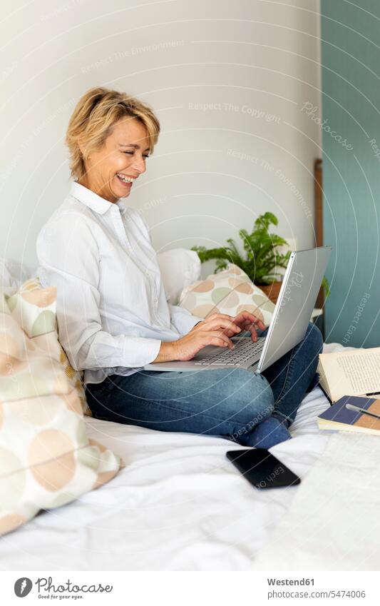 Glückliche reife Frau sitzt zu Hause am Bett und benutzt einen Laptop Leute Menschen People Person Personen Alleinstehende Alleinstehender Singles