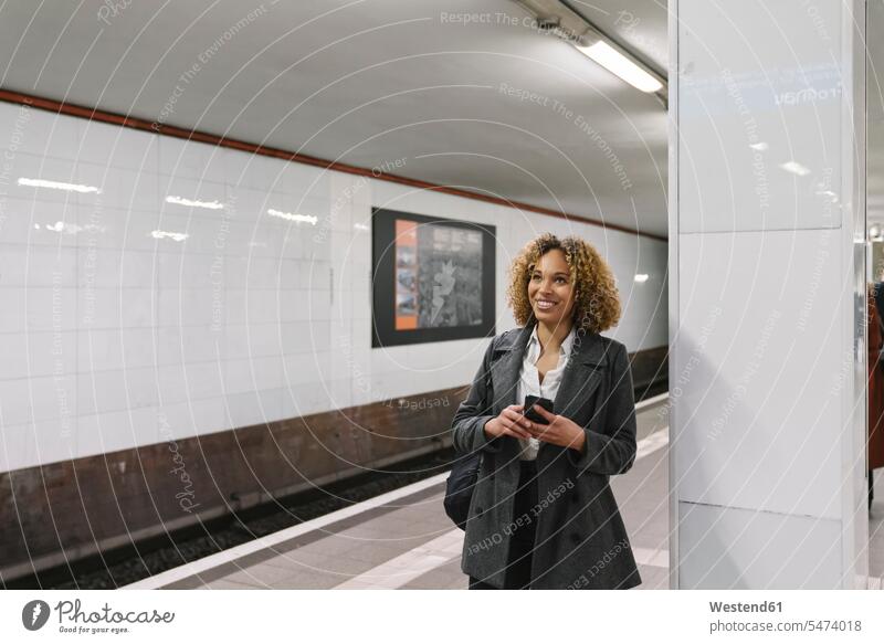 Lächelnde Frau mit Handy wartet in U-Bahn-Station geschäftlich Geschäftsleben Geschäftswelt Geschäftsperson Geschäftspersonen Businessfrau Businessfrauen