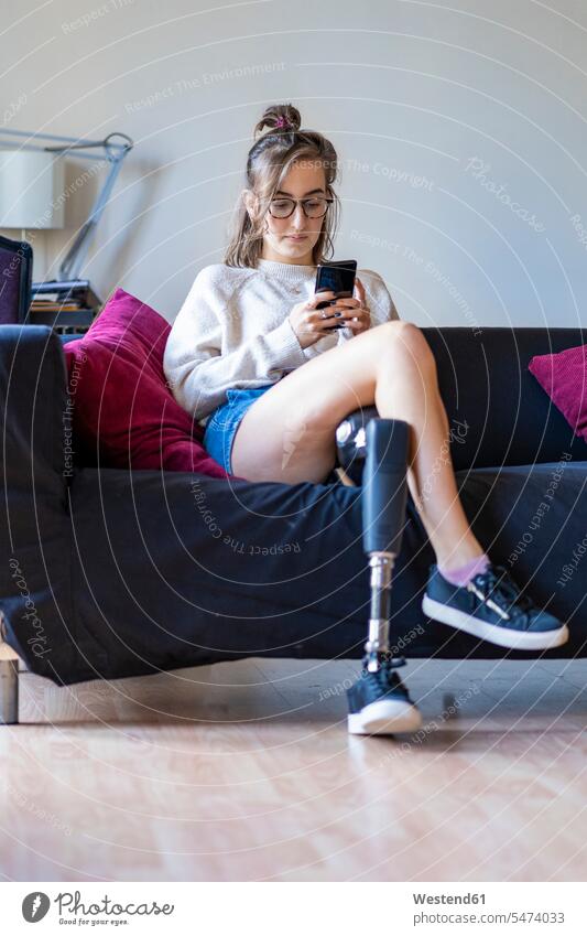 Junge Frau mit Beinprothese sitzt zu Hause auf dem Sofa und benutzt ein Smartphone Leute Menschen People Person Personen Alleinstehende Alleinstehender Singles