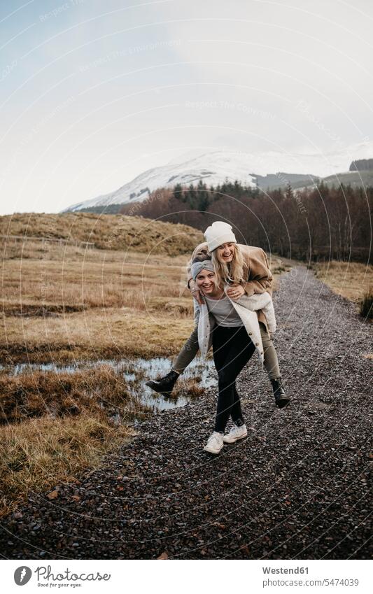 Großbritannien, Schottland, glückliche junge Frau trägt Freund Huckepack in ländlicher Landschaft Freundinnen Glück glücklich sein glücklichsein weiblich Frauen
