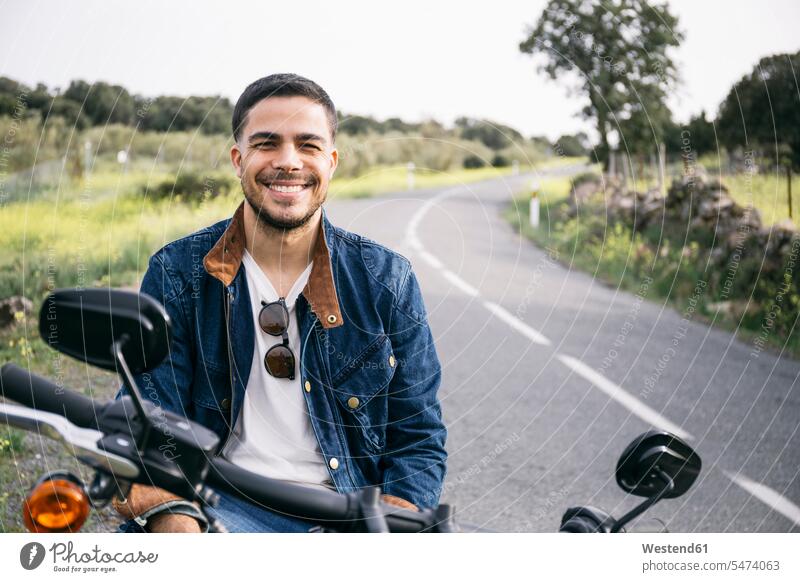 Lächelnder junger Biker sitzt auf einem Motorrad auf dem Land Farbaufnahme Farbe Farbfoto Farbphoto Spanien Freizeitbeschäftigung Muße Zeit Zeit haben