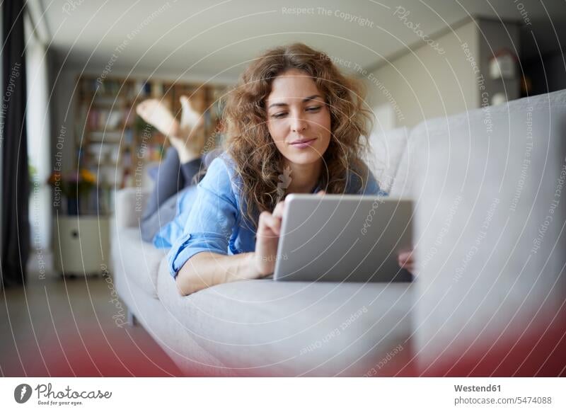 Schöne Frau benutzt digitales Tablet, während sie zu Hause auf dem Sofa liegt Farbaufnahme Farbe Farbfoto Farbphoto Innenaufnahme Innenaufnahmen innen drinnen