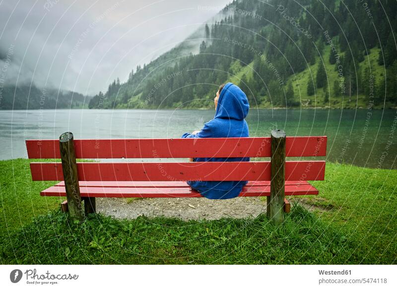 Frau mit blauer Kapuze sitzt auf Bank am Seeufer und schaut auf Berge Farbaufnahme Farbe Farbfoto Farbphoto Österreich Außenaufnahme außen draußen im Freien Tag