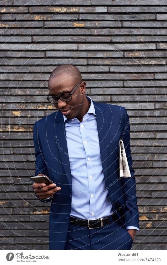 Porträt eines lächelnden Geschäftsmannes mit Zeitung, der eine Brille und einen blauen Anzug trägt und auf ein Mobiltelefon schaut Businessmann Businessmänner