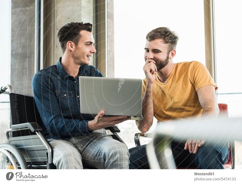 Junger Geschäftsmann im Rollstuhl zeigt Laptop einem Kollegen im Büro Arbeitskollege Arbeitskollegen Partner Partnerschaften Job Berufe Berufstätigkeit