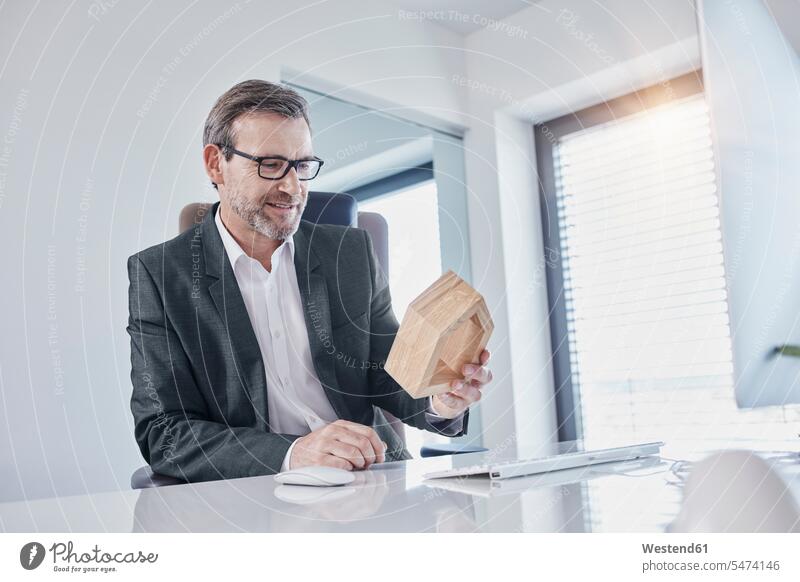 Lächelnder Geschäftsmann am Schreibtisch im Büro beim Betrachten eines Architekturmodells Arbeitstisch Schreibtische Office Büros lächeln ansehen Businessmann