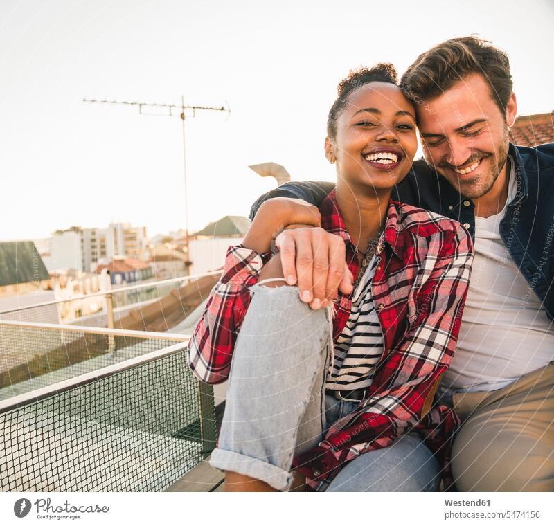 Glückliches, liebevolles junges Paar sitzt abends auf dem Dach entspannen relaxen knuddeln schmusen sitzend Arm umlegen Umarmung Umarmungen Jahreszeiten