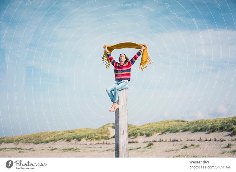 Reife Frau sitzt auf einem Holzpfahl am Strand, hält Schal in den Wind Schals Beach Straende Strände Beaches Brisen Winde windig Holzpfosten flattern sitzen