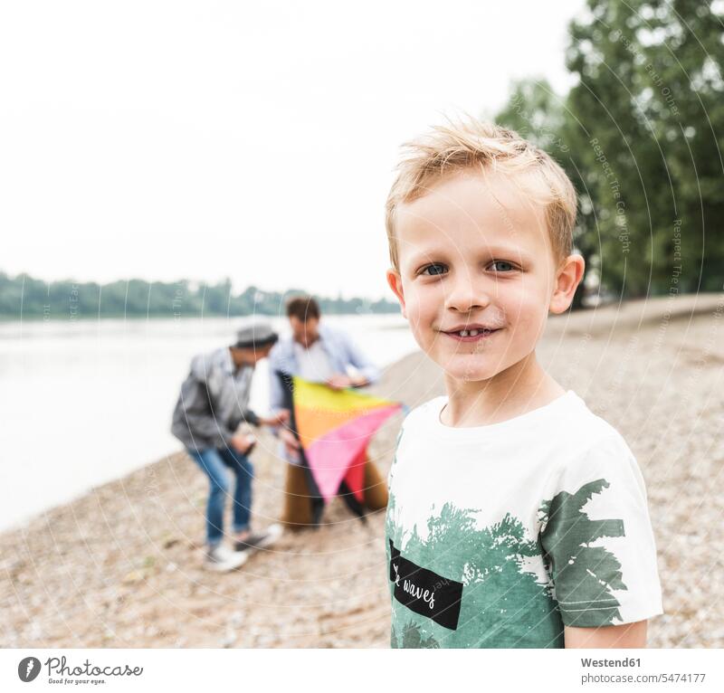 Porträt eines lächelnden Jungen mit Vater und Bruder, die einen Drachen am Flussufer halten Sohn Söhne Papas Väter Vati Vatis Papis Portrait Porträts Portraits
