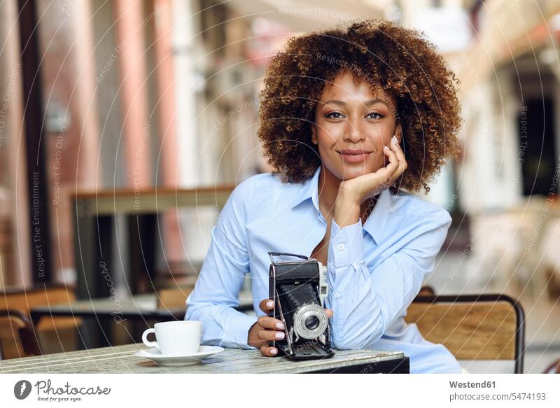 Porträt einer lächelnden Frau mit Oldtimer-Kamera, die in einem Café im Freien sitzt Cafe Kaffeehaus Bistro Cafes Cafés Kaffeehäuser weiblich Frauen Portrait
