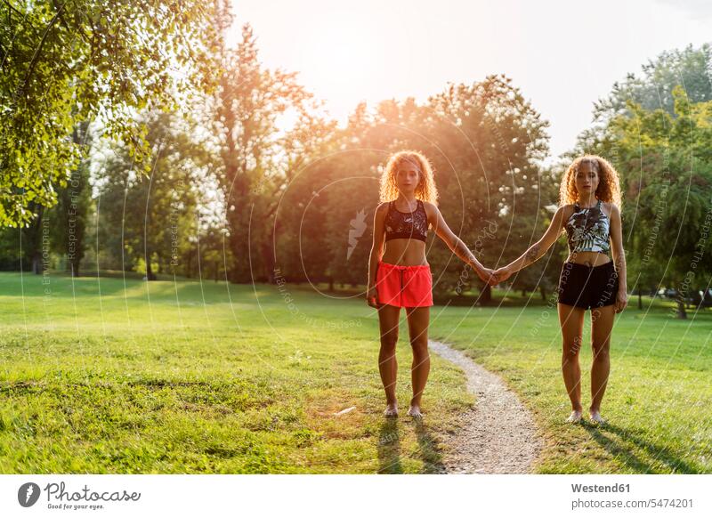 Zwillingsschwestern stehen nebeneinander in einem Park bei Gegenlicht halten Hände Parkanlagen Parks stehend steht Seite an Seite Gegenlichtaufnahme