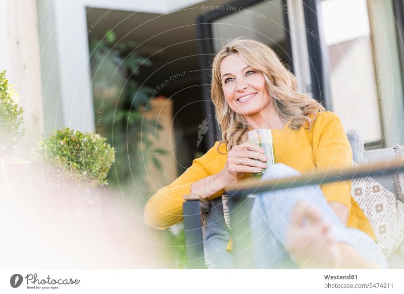 Porträt einer lächelnden reifen Frau, die mit grünem Smoothie auf der Terrasse sitzt Gläser Trinkglas Trinkgläser Stuehle Stühle entspannen relaxen sitzend