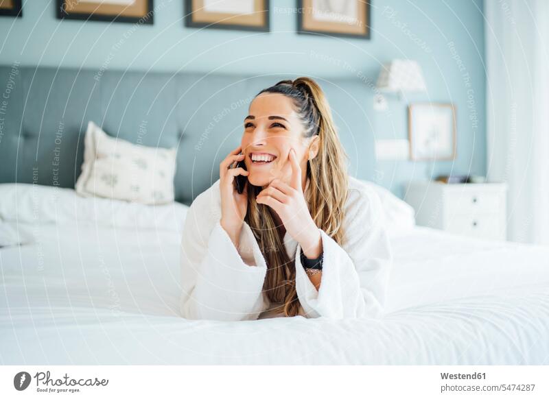 Fröhliche Frau mittleren Alters, die im Hotel auf dem Bett liegt und über ein Smartphone telefoniert Farbaufnahme Farbe Farbfoto Farbphoto Spanien