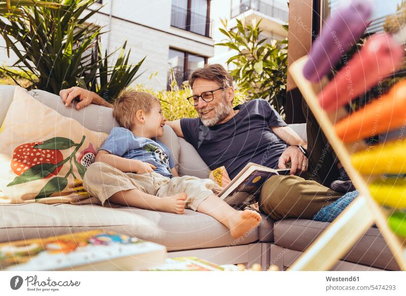 Vater und Sohn lesen ein Buch auf ihrer Terrasse T-Shirt Sofa lächeln Sommer entspannt Freude fröhlich Geborgenheit genießen glücklich Liebe Zufriedenheit