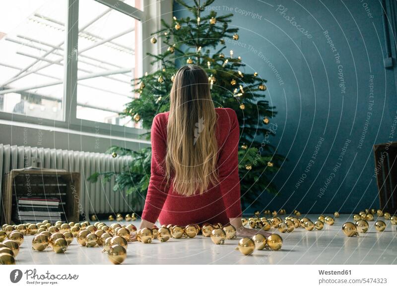 Rückenansicht einer Frau, die mit vielen goldenen Weihnachtskugeln auf dem Boden sitzt und auf den Weihnachtsbaum schaut sitzen sitzend Fußboden Fußboeden