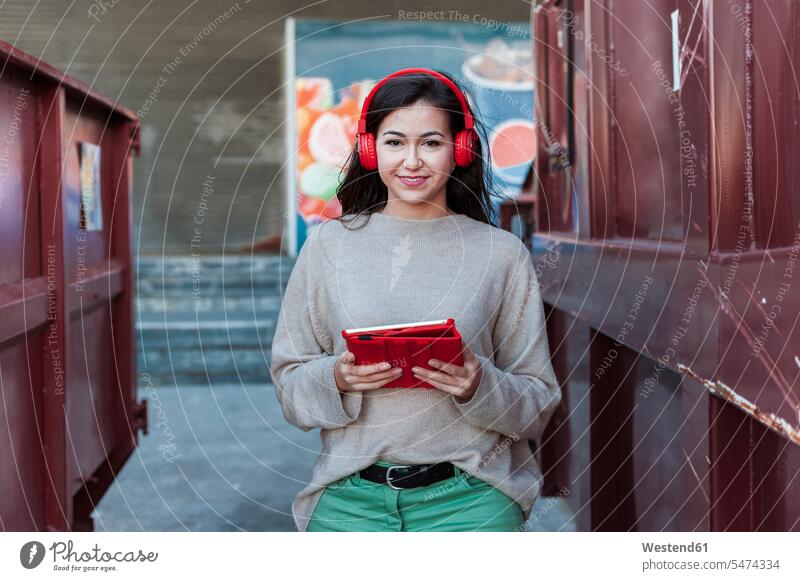 Lächelnde schöne Frau hört Musik über Kopfhörer, während sie ein digitales Tablet hält Farbaufnahme Farbe Farbfoto Farbphoto Außenaufnahme außen draußen