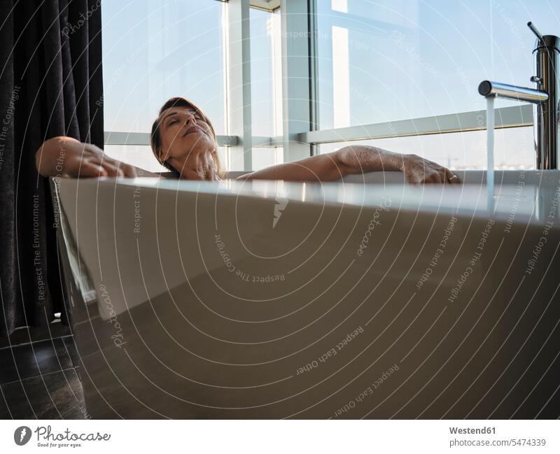 Entspannte ältere Frau nimmt Bad in Badewanne gegen Fenster im Luxus-Hotelzimmer Farbaufnahme Farbe Farbfoto Farbphoto Innenaufnahme Innenaufnahmen innen