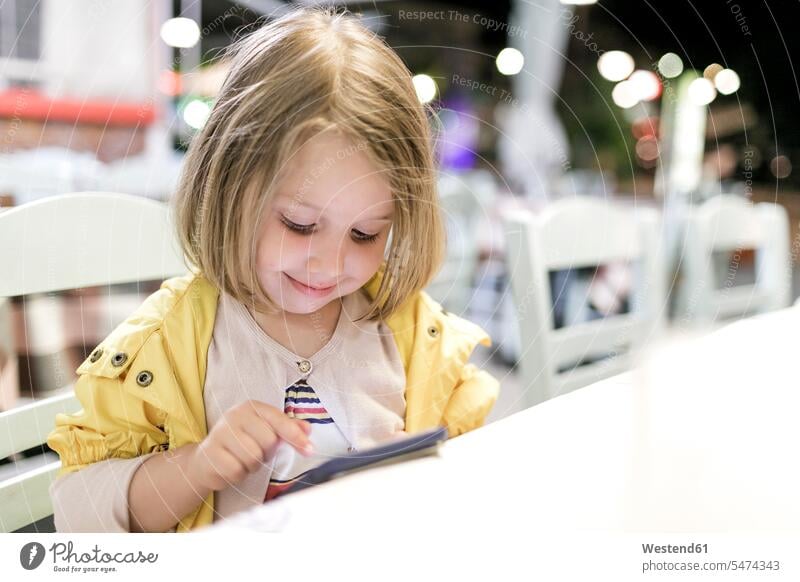 Porträt eines lächelnden kleinen Mädchens, das in einem Restaurant sitzt und mit einem Smartphone spielt weiblich spielen Lokal Speiserestaurant Lokale