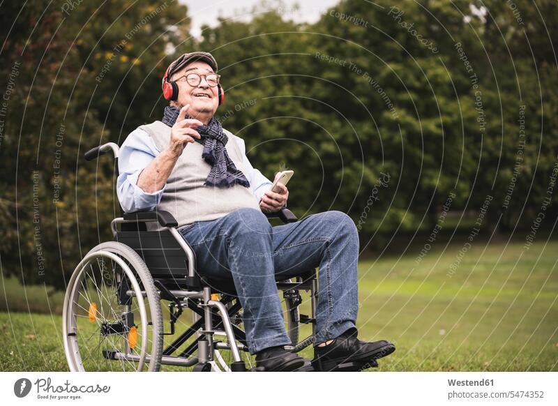 Porträt eines glücklichen älteren Mannes, der im Rollstuhl sitzt und mit Smartphone und Kopfhörern Musik hört Leute Menschen People Person Personen Europäisch