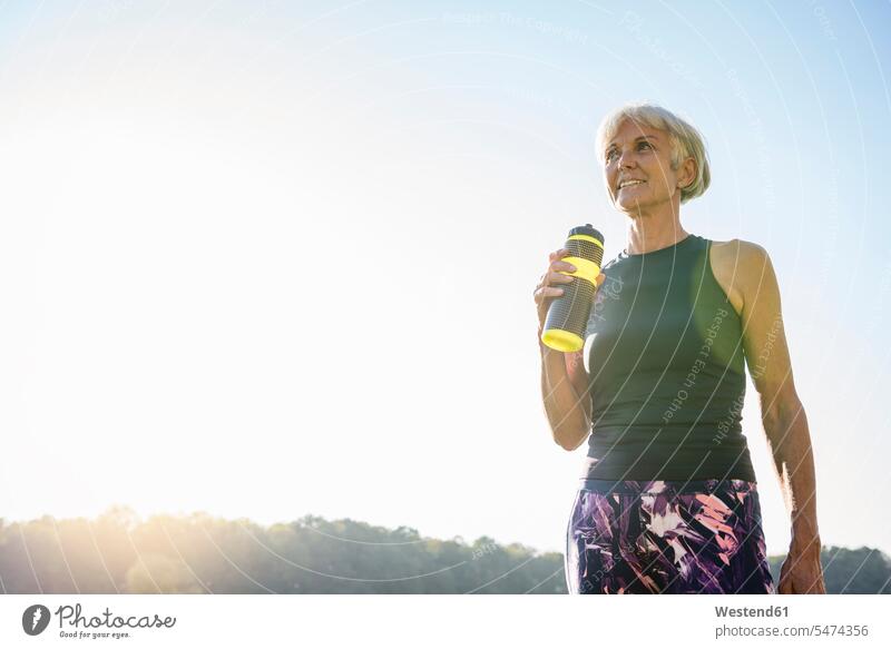 Lächelnde sportliche ältere Frau hält Flasche unter blauem Himmel Flaschen halten lächeln weiblich Frauen Seniorin Seniorinnen Erwachsener erwachsen Mensch