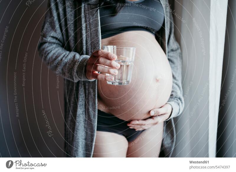 Schwangere Frau hält Trinkglas in der Hand, während sie zu Hause den Bauch berührt Farbaufnahme Farbe Farbfoto Farbphoto Innenaufnahme Innenaufnahmen innen