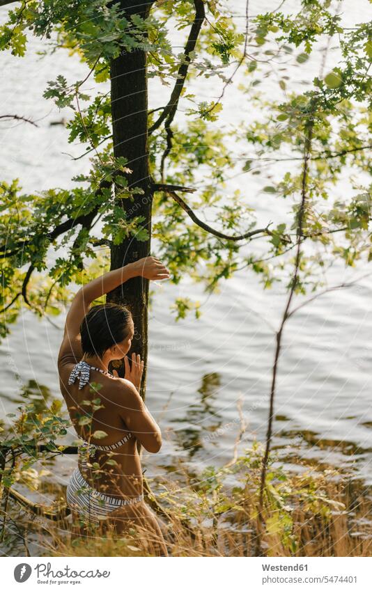 Rückansicht einer Frau im Bikini am Baumstamm an einem See Stamm Stämme Baumstämme Seen weiblich Frauen Bikinis Bäume Baeume Gewässer Wasser Erwachsener