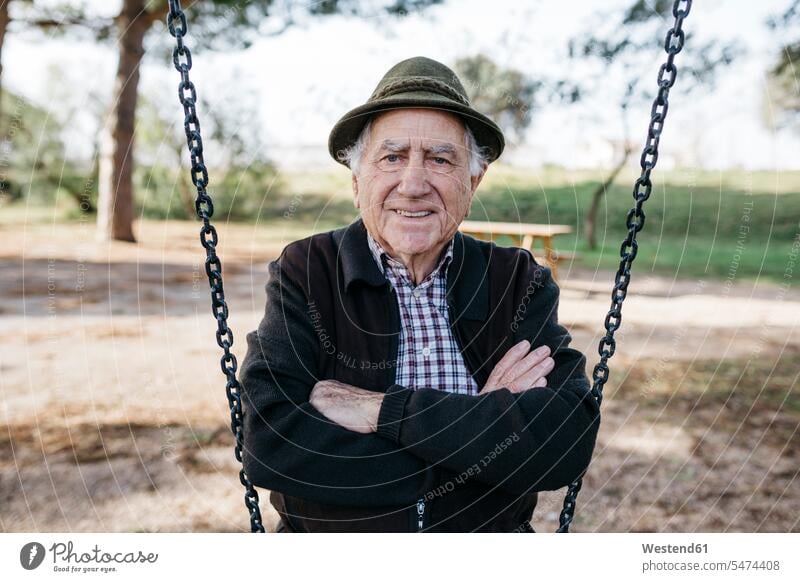 Alter Mann auf Schaukel im Park sitzend, mit verschränkten Armen Hüte Schaukeln freuen Glück glücklich sein glücklichsein Pension pensioniert Pensionierung