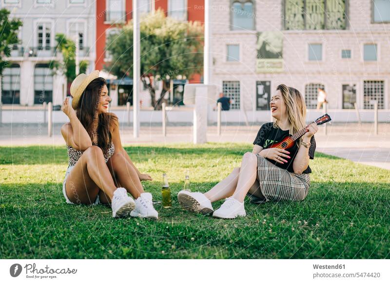 Fröhliche Frau spielt Ukulele, während sie mit einer Freundin auf Grasland sitzt Farbaufnahme Farbe Farbfoto Farbphoto Portugal Freizeitbeschäftigung Muße Zeit