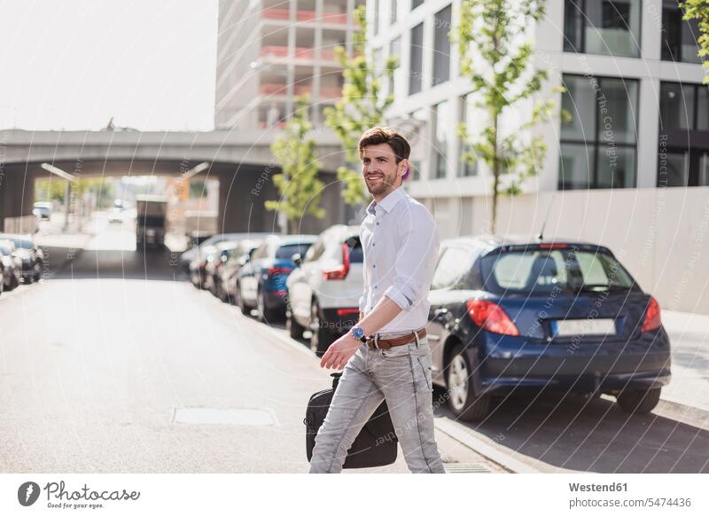 Lächelnde Geschäftsmann mit Laptop-Tasche in der Stadt die Straße überqueren Laptoptasche staedtisch städtisch unterwegs auf Achse in Bewegung Strassen Straßen