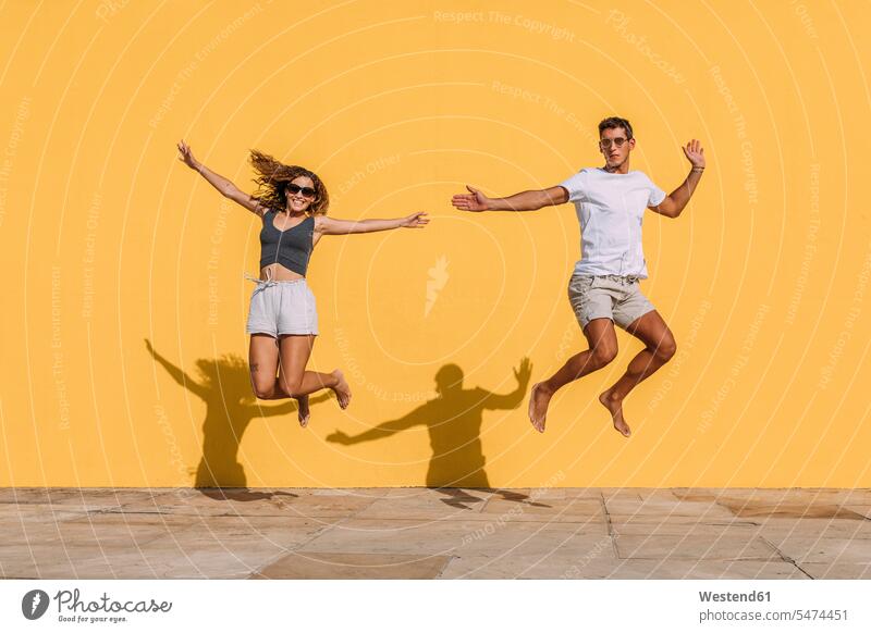 Junges Paar springt vor eine gelbe Mauer T-Shirts Brillen Sonnenbrillen springen Spruenge Sprünge hüpfen Jahreszeiten sommerlich Sommerzeit freuen gefühlvoll