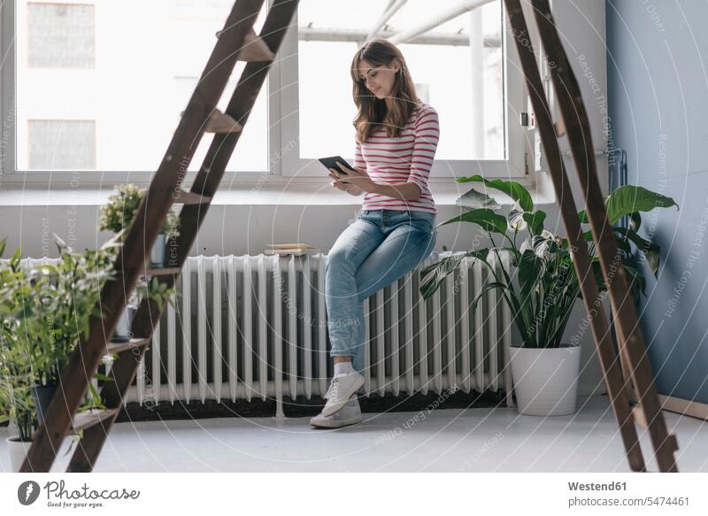 Frau sitzt in ihrem neuen Zuhause am Heizkörper, liest E-Book, umgeben von Pflanzen lesen Lektüre weiblich Frauen eBook zu Hause daheim Topfpflanze Topfpflanzen