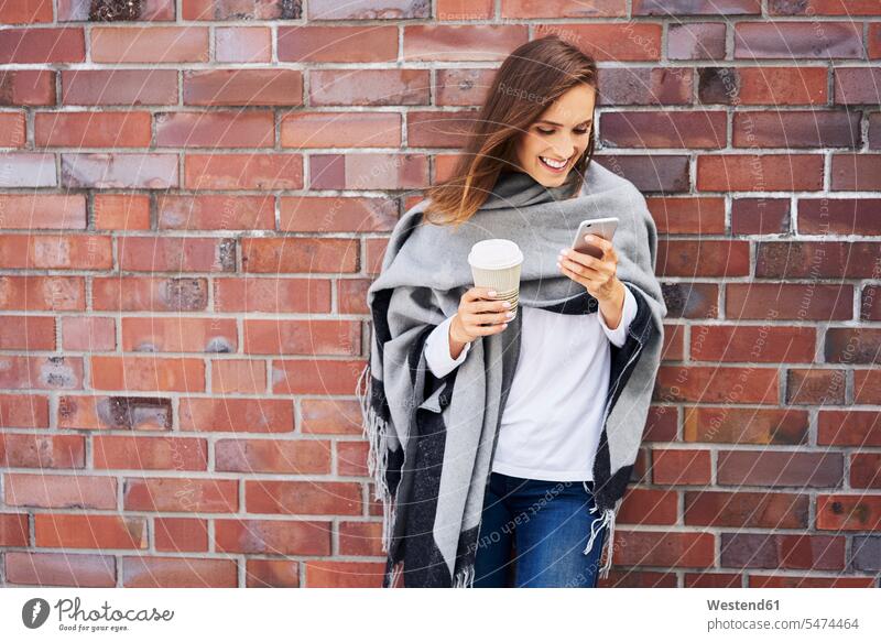 Glückliche Frau mit Kaffee zum Handy vor der Backsteinmauer Coffee to go zum mitnehmen Backsteinwand Backsteinmauern Smartphone iPhone Smartphones weiblich