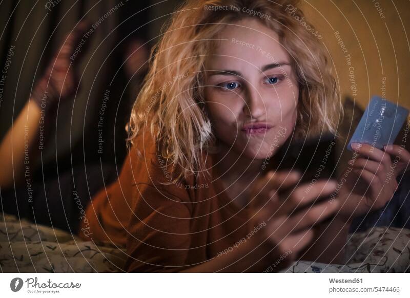 Porträt einer blonden jungen Frau, die zu Hause ein Smartphone und eine Kreditkarte benutzt EC-Karte Kreditkarten Betten Telekommunikation Handies Handys