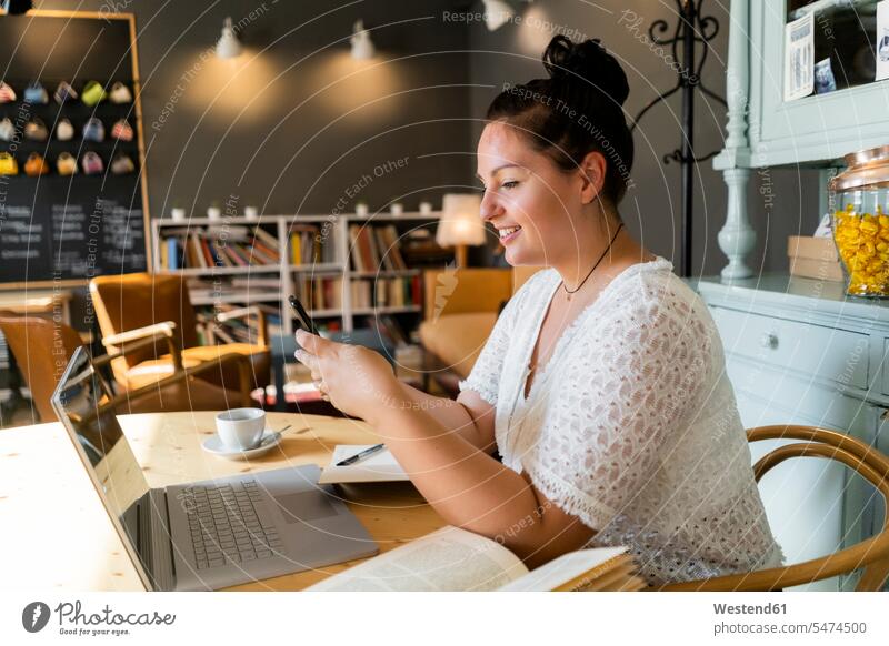 Junge Frau mit Büchern und Laptop auf dem Tisch mit einem Smartphone im Café Farbaufnahme Farbe Farbfoto Farbphoto Innenaufnahme Innenaufnahmen innen drinnen