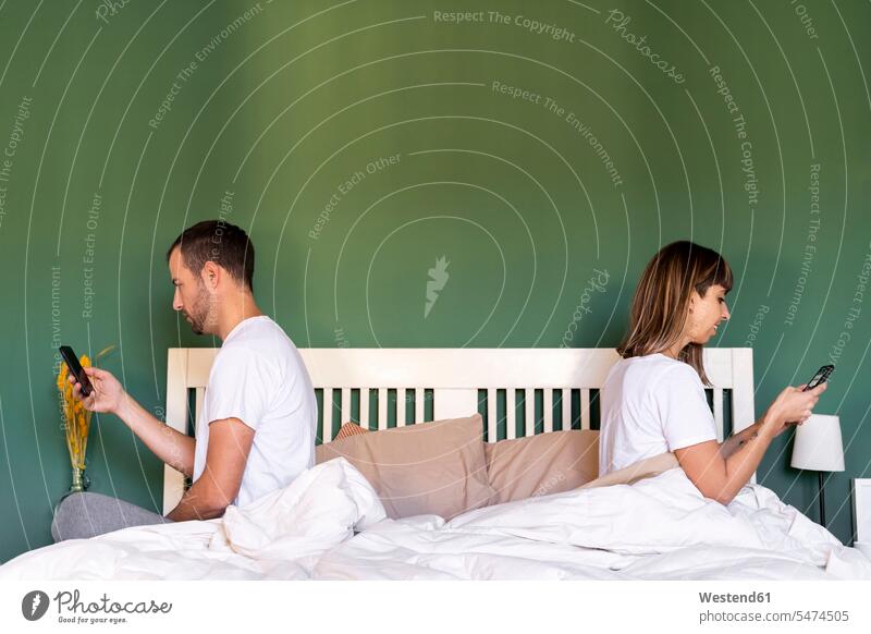 Mid adult couple using mobile phone while sitting on bed at home Farbaufnahme Farbe Farbfoto Farbphoto Innenaufnahme Innenaufnahmen innen drinnen morgens Morgen