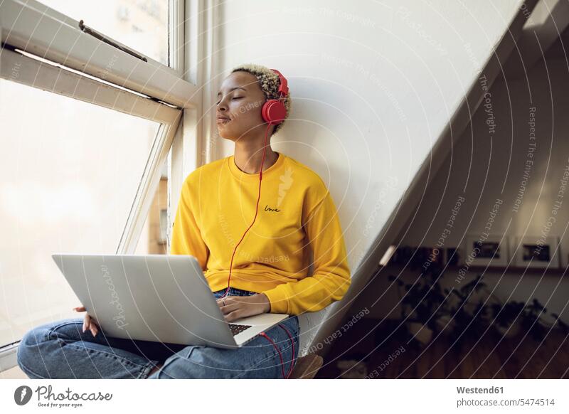 Junge Frau sitzt zu Hause am Fenster mit Laptop und Kopfhörern Hochschueler Hochschüler Studenten Studierende Studierender Leute Menschen People Person Personen