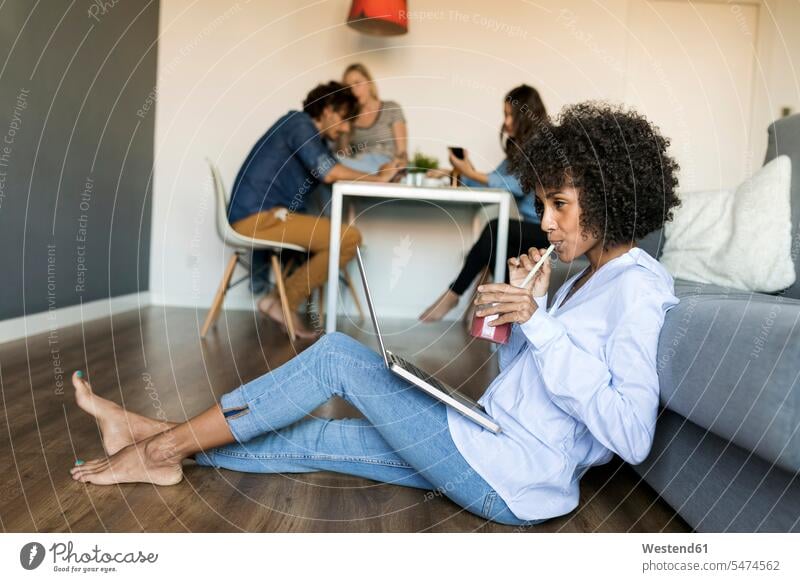 Frau mit Softdrink sitzt auf dem Boden mit Laptop mit Freunden im Hintergrund Böden Boeden weiblich Frauen Erfrischungsgetränk Erfrischungsgetraenk sitzen