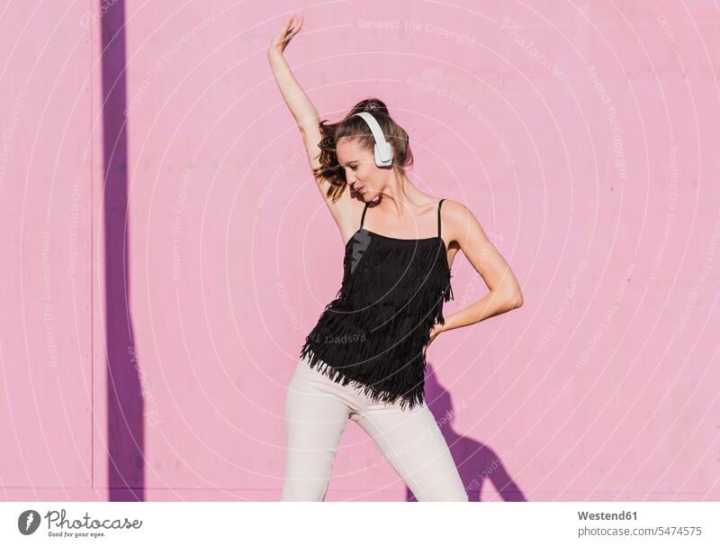 Glückliche junge Frau mit Kopfhörer tanzen vor rosa Wand Kopfhoerer glücklich glücklich sein glücklichsein tanzend weiblich Frauen rosafarben Wände Waende