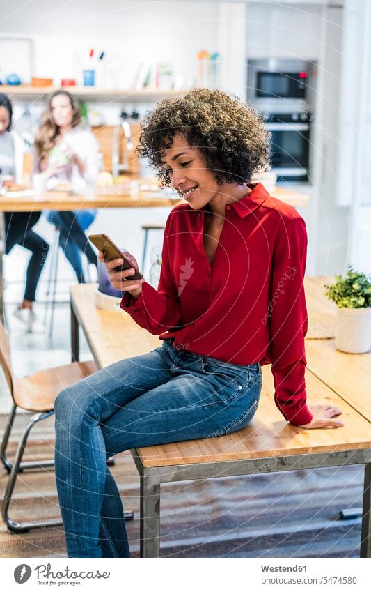 Lächelnde Frau mit Handy sitzt auf dem Tisch sitzen sitzend lächeln Mobiltelefon Handies Handys Mobiltelefone weiblich Frauen Tische Telefon telefonieren