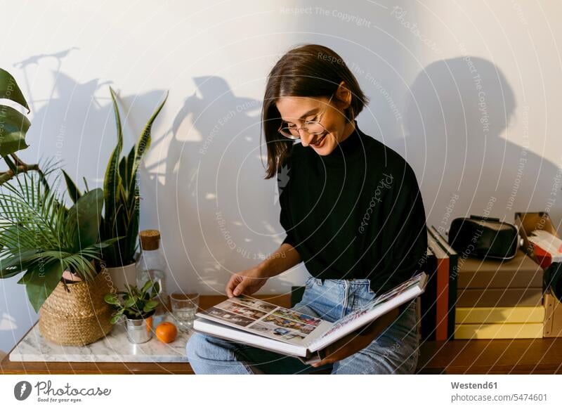 Lächelnde Frau schaut Fotos im Fotoalbum an, während sie zu Hause sitzt Farbaufnahme Farbe Farbfoto Farbphoto Innenaufnahme Innenaufnahmen innen drinnen