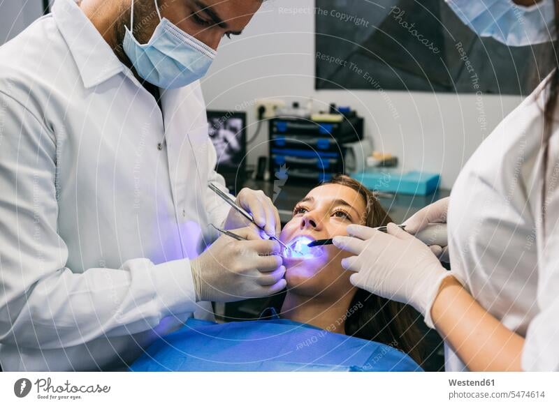 Männlicher Zahnarzt mit Maske und Handschuhen untersucht Zähne einer Patientin mit Hilfe einer Assistentin in der Klinik Farbaufnahme Farbe Farbfoto Farbphoto