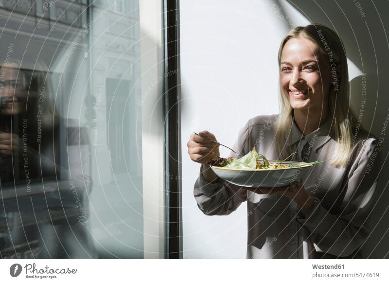Lächelnde junge Frau steht am Fenster in der Sonne und isst einen Salat blond blonde Haare blondes Haar Gesunde Ernährung Ernaehrung Gesunde Ernaehrung